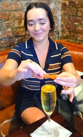 Person in striped dress twisting an orange peel over a sparkling champagne flute at a cozy brick-walled bar with a leather banquette