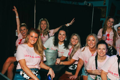 Group of smiling women in matching pink “Let’s Go Girls” shirts posing with drinks and a metal bucket during a lively bachelorette girls’ night out in a dimly lit bar