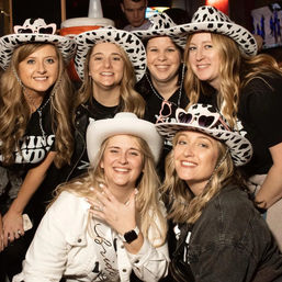 Smiling friends at a bachelorette party in a bar, wearing cow-print cowboy hats and sunglasses, bride showing engagement ring