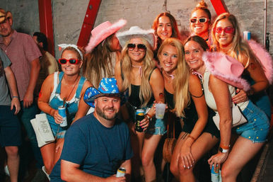 Smiling group of friends at an indoor party wearing cowboy hats and pink feathered accessories, sunglasses and denim outfits, holding drinks in front of a rustic brick wall.