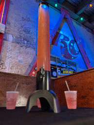 Neon-lit bar interior with graffiti-covered walls and a tall pink drink tower on a table flanked by two plastic cups with straws — lively nightlife scene.