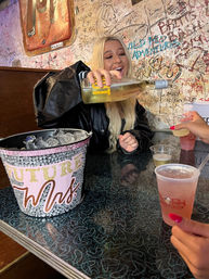 Blonde woman laughing as she pours white wine from a bottle into plastic cups at a graffiti-covered bar booth, ice bucket reading Future Mrs., friends holding cocktails — lively bachelorette celebration.