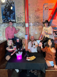 Six friends toasting drinks in a lively graffiti-covered bar booth with a neon-lit drink tower, bucket of beers, chips and salsa, and one guest wearing a cowboy hat.