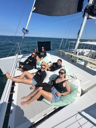 Group of friends lounging on a catamaran trampoline and mint cushions, wearing sunglasses and summer clothes, enjoying a sunny sailing day on the calm blue ocean.