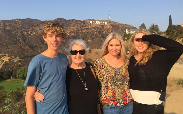 Multigenerational group of four posing on a sunny hillside trail with the Hollywood sign on Mount Lee and the Hollywood Hills skyline in Los Angeles.