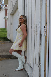 Outdoor portrait of a smiling woman in a yellow polka-dot dress and white knee-high boots leaning against a weathered white barn door at a farmhouse