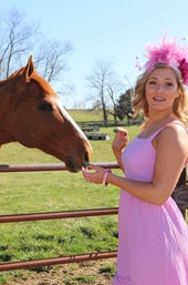 Woman in a pink sundress and feathered headpiece feeding a chestnut horse over a metal fence in a sunny green pasture with bare trees and farm fencing in the background.