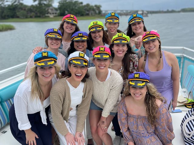 Smiling group of women in colorful captain caps posing on a boat near a coastal inlet, enjoying a daytime waterfront celebration.