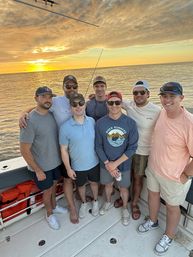 Seven friends posing on a boat deck during a golden ocean sunset, wearing casual summer clothes, hats and sunglasses on a relaxed coastal fishing trip.