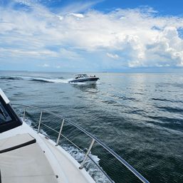 Bow of a white motorboat in the foreground cutting through calm open ocean waters while a nearby speedboat zips by under blue sky and puffy clouds