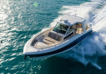 Aerial view of a white luxury motorboat cutting through turquoise coastal water with a sunlit bow lounge, hardtop cabin, and frothy wake trailing behind.