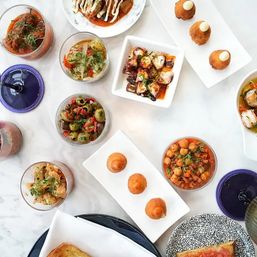 Overhead shot of a colorful Spanish tapas spread on a marble table — marinated olives, chickpea stew, grilled octopus, crispy croquettes and tomato-topped toast in small glass bowls and plates.