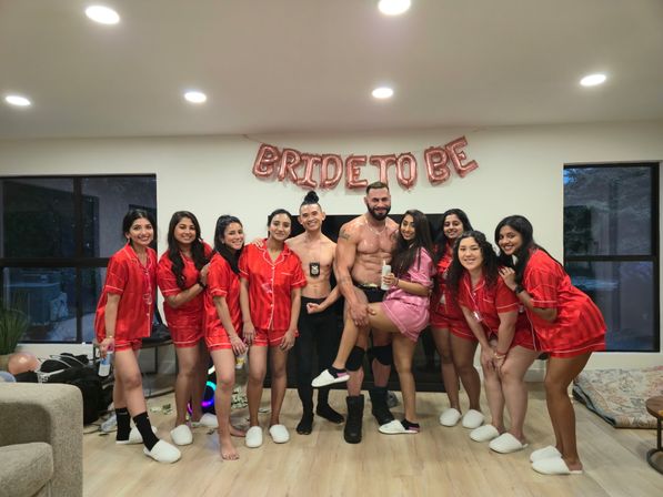 Indoor bachelorette party group photo: women in matching red pajamas and one in pink pose with two male entertainers under a rose-gold "BRIDE TO BE" banner.