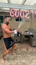 Shirtless man in a short black apron popping a champagne bottle on a backyard patio, spray arcing beneath rose-gold letter balloons with wicker dining furniture and tiled pavers in the background — playful outdoor celebration.