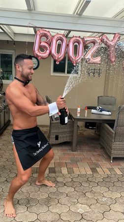 Shirtless man in a short black apron popping a champagne bottle on a backyard patio, spray arcing beneath rose-gold letter balloons with wicker dining furniture and tiled pavers in the background — playful outdoor celebration.
