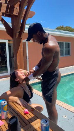 Sunny backyard pool scene: a shirtless man in a cap and striped apron leans over a wooden table under a pergola, pouring a drink into a laughing woman in a black bikini beside the pool, canned drinks on the table.