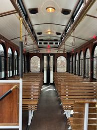 Sunlit vintage trolley interior with rows of wooden bench seats, arched windows, brass poles and leather hand straps flanking a central aisle
