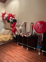 Bedroom decorated with red, black and white balloon garland with shiny red star accents over a bed; large silver letter balloons tied to a dresser and a big red monogram balloon on a hardwood floor.