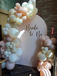 Cheerful pastel balloon arch in cream and blush framing a round backdrop with gold "Bride to Be" script at an indoor bridal shower venue with exposed brick wall.