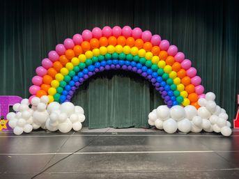 Playful rainbow balloon arch of pink, orange, yellow, green, blue and purple rows with white balloon cloud bases on a stage in front of dark green theater curtains — festive balloon arch event decor backdrop