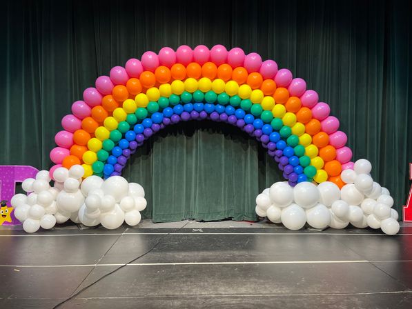 Playful rainbow balloon arch of pink, orange, yellow, green, blue and purple rows with white balloon cloud bases on a stage in front of dark green theater curtains — festive balloon arch event decor backdrop