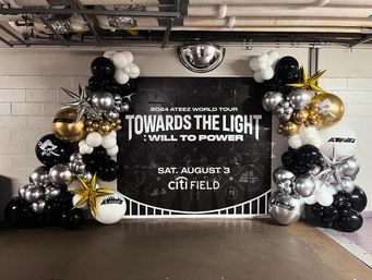 Stadium corridor display: black 'TOWARDS THE LIGHT' tour backdrop framed by festive gold, silver, black and white balloon garlands with star-shaped foil balloons.