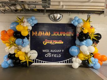 Colorful balloon arch framing a large concert poster for classic rock bands with bold logos and an event date, displayed in a stadium concourse.