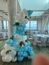 Playful sky-blue and white balloon garland with eucalyptus wrapped around a mosaic pillar in an ocean-view banquet hall set for an event.