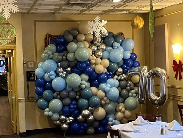 Indoor party scene with a large organic balloon wall in shades of blue, gray and metallic silver, topped by a white snowflake and flanked by silver “70” foil number balloons next to a set dining table.