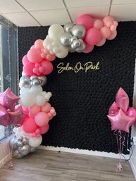 Indoor salon photo backdrop featuring a black rose-textured wall with neon script, pink, white and silver balloon arch and pink star-shaped foil balloons