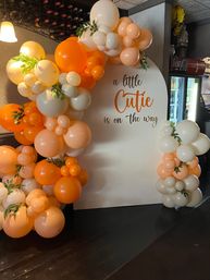 Peach, orange and cream balloon arch with greenery framing a white backdrop reading 'a little Cutie is on the way' — baby shower photo backdrop and event decor.
