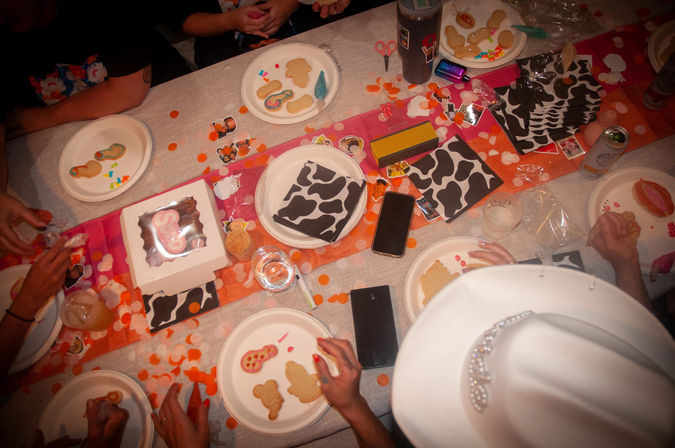 Overhead view of a lively cookie-decorating party table with paper plates holding shaped cookies, icing and sprinkles, orange confetti, cow-print napkins, drinks and phones, and a white cowboy hat in the foreground.
