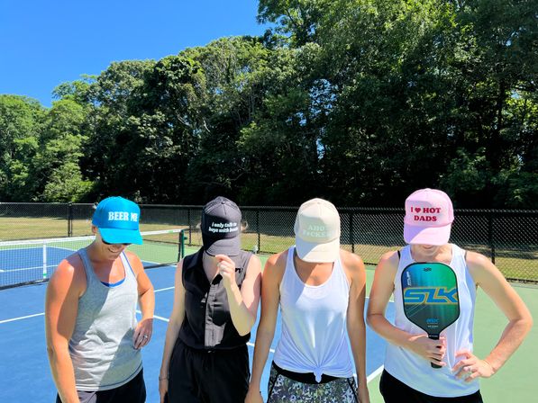 Four people on an outdoor pickleball court on a sunny day, heads bowed and wearing colorful caps with playful slogans while one player holds a pickleball paddle; trees and a chain-link fence frame the background.