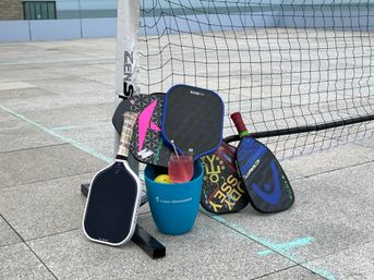 Assorted colorful pickleball paddles and balls propped against a net on a rooftop waterfront outdoor court, a blue bucket holds a yellow ball and a pink cocktail in a stemmed glass on the concrete surface with ocean visible beyond the court.