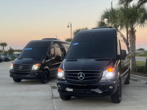 Two black passenger vans parked in a palm-lined parking lot at sunset, headlights and a blue "LIMO" display glowing on a windshield.