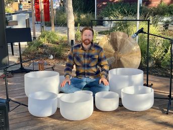Person seated cross-legged on an outdoor wooden stage surrounded by white crystal singing bowls and a large bronze gong, with microphones and stands set up for a sound-healing performance.