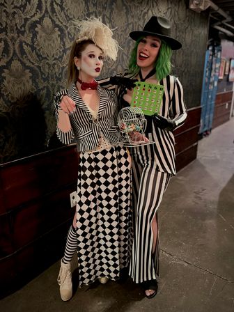 Two people in black-and-white carnival costumes pose in a dim hallway against damask wallpaper — one with white face paint, feathered headpiece and harlequin check skirt; the other with a green wig, fedora, striped suit holding a bingo card and a small bingo cage with colorful balls.