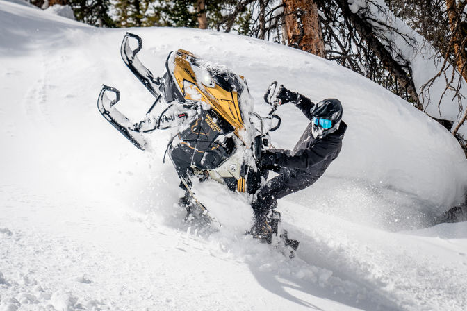 Snowmobiler in black gear and blue goggles lifts the front of a yellow snowmobile through deep powder near snow-covered trees in a mountain forest