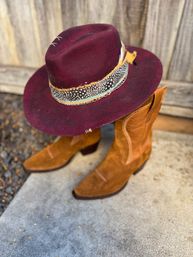 Burgundy felt wide-brim western hat with a feather-and-bead hatband resting on tan suede cowboy boots against a rustic wooden fence