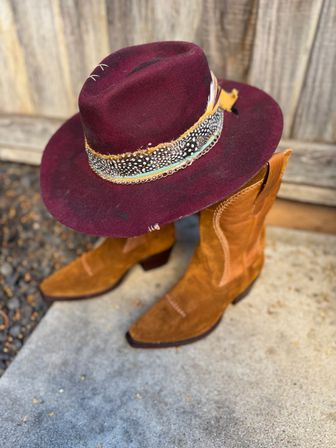 Burgundy felt western hat with feather-and-bead hatband perched on tan suede cowboy boots against a weathered wood fence, rustic western style