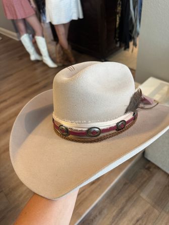 Beige felt cowboy hat with braided leather and concho-studded band and feather accent, held over a hardwood floor with blurred cowgirl boots and sundresses in the background.