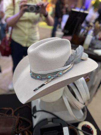Light gray felt cowboy hat with feather and turquoise concho band displayed on a mannequin at a busy artisan market stall