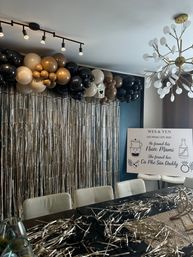 Las Vegas party decor with black, gold and cream balloon garland over a silver fringe backdrop, dining table scattered with metallic streamers, a modern chandelier, and a personalized couple sign.