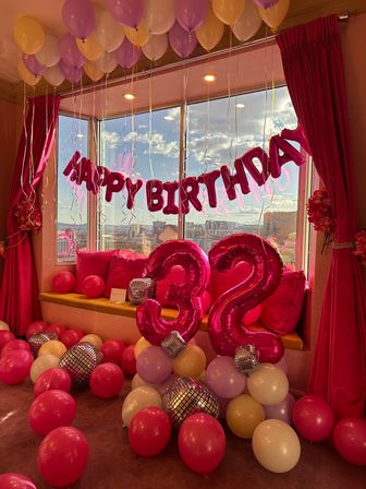 Bright pink birthday setup with foil "HAPPY BIRTHDAY" banner and large metallic '32' balloons surrounded by pink, purple and cream balloons and mirrored disco-ball accents on a window seat overlooking a city skyline.