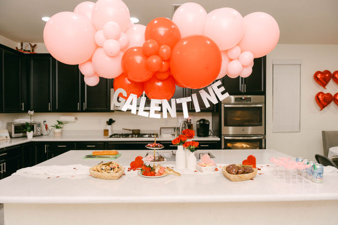 Festive Galentine's brunch setup on a white marble kitchen island with oversized pink and red balloons and a silver 'GALENTINE' banner overhead, plates of pastries, strawberries, chocolates, red flowers and drink glasses.