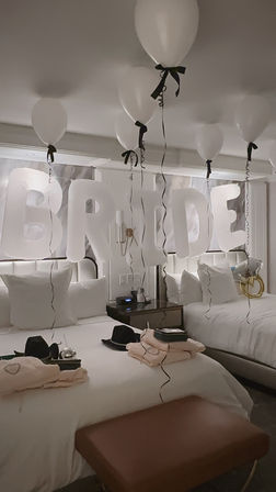 Bridal suite hotel room decorated for a bachelorette with large white letter balloons spelling BRIDE and floating white balloons tied with black ribbons above two neatly made beds.