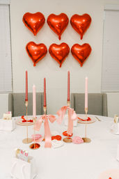 Valentine’s Day indoor table setup with six red heart foil balloons on a white wall, gold candle holders holding pink and red taper candles tied with pink satin bows, woven heart decorations and small white gift bags.