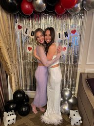 Two women hugging at an indoor bachelorette photo booth — bride in white sequined gown with veil and feather hem, friend in lavender sequin two-piece, silver fringe backdrop, black/red/silver balloons and playing-card decorations.
