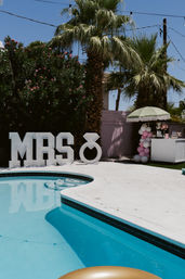 Sunlit backyard pool scene with large light-up "MRS" marquee and ring prop, palm trees, pastel umbrella, pink-and-white balloon column and dessert cart for a bridal shower