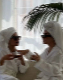 Two people in white bathrobes and towel turbans wearing sunglasses, sipping coffee in a bright spa-like resort lounge under tropical palm fronds.